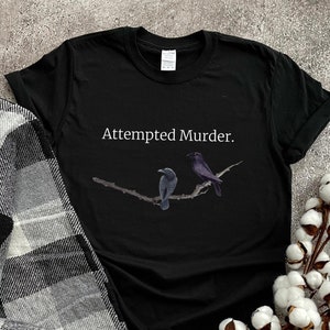 Puede incluir: Camiseta negra con el texto "Attempted Murder." y un gr&aacute;fico de dos p&aacute;jaros negros en una rama.