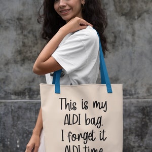 Op de afbeelding: Een witte canvas tote bag met blauwe handvatten. De tas heeft zwarte tekst die "This is my ALDI bag. I forget it ALDI time." zegt.