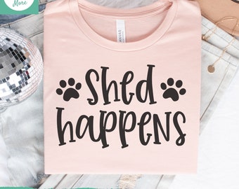 May include: A pink t-shirt with the text "Shed happens" in black lettering. Two black paw prints are on either side of the text.