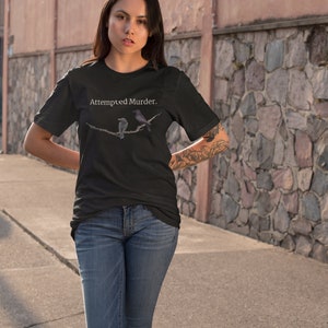 Puede incluir: Una camiseta negra con el texto "Attempted Murder." impreso en blanco. La camiseta presenta un gr&aacute;fico de dos p&aacute;jaros en una rama.