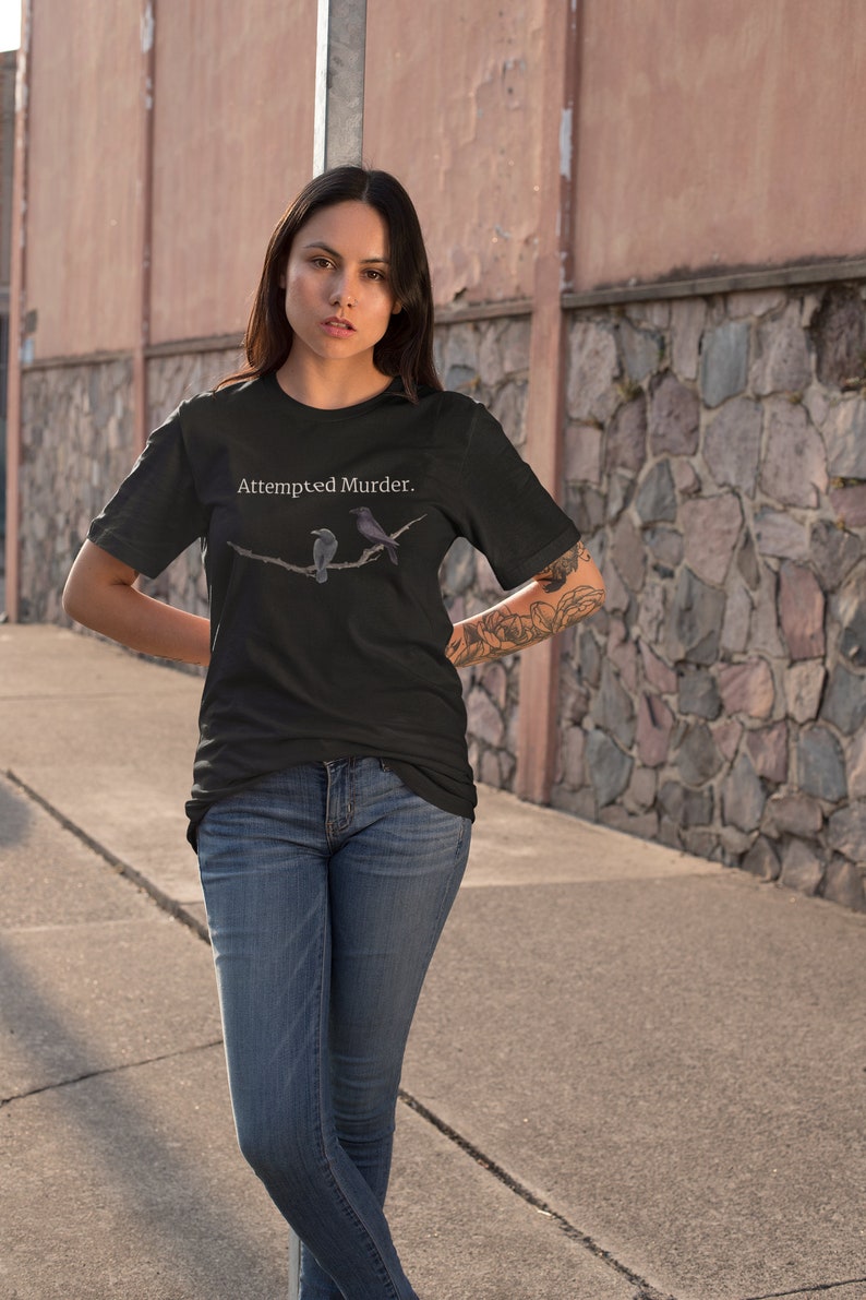 Puede incluir: Una camiseta negra con el texto "Attempted Murder." impreso en blanco. La camiseta presenta un gr&aacute;fico de dos p&aacute;jaros en una rama.