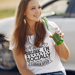 May include: A woman wearing a white t-shirt with the text "I'm not drunk, I'm socially lubricated" printed on it. She is holding a green glass bottle and smiling.