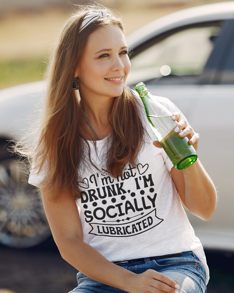 May include: A woman wearing a white t-shirt with the text "I'm not drunk, I'm socially lubricated" printed on it. She is holding a green glass bottle and smiling.