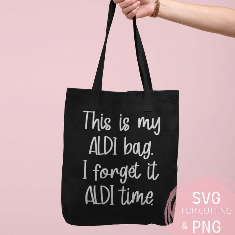 Op de afbeelding: Een zwarte tote bag met de tekst "This is my ALDI bag. I forget it ALDI time." in witte letters. De tas wordt vastgehouden door een hand tegen een roze achtergrond. De tekst "SVG FOR CUTTING & PNG" is zichtbaar in de rechterbenedenhoek van de afbeelding.