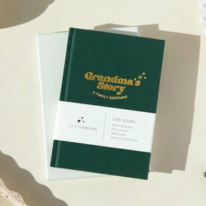 May include: A green journal with gold lettering that reads "Grandma's Story A Family Keepsake". The journal has a white label with the text "LIFE STORY Share Memories Life Lessons Reflections Notes to Loved Ones" and the text "CATCHYOURDREAMS" in gold lettering.