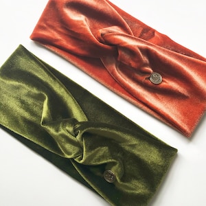Op de afbeelding: Twee fluwelen hoofdbanden, een in een diepe groene kleur en de andere in een verbrand oranje. Beide hoofdbanden hebben een kleine, ronde metalen knop aan de zijkant.