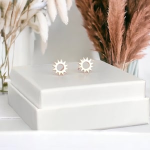 Könnte beinhalten: Ein Paar goldene Sonnenohrringe in einer weißen Schachtel. Die Ohrringe sind klein und zart, mit einem schlichten Design.
