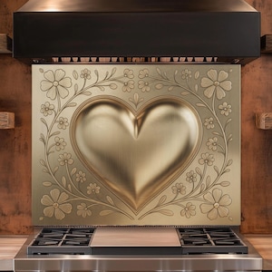 May include: A gold-colored kitchen backsplash with a heart-shaped design, surrounded by floral patterns. The backsplash is installed above a stainless steel gas range. The image also shows a black range hood and wooden shelves.