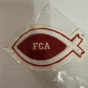 Könnte beinhalten: Ein rot-weißer Aufnäher in Form eines Fisches mit den Buchstaben "FCA" in Weiß. Der Aufnäher befindet sich in einer durchsichtigen Plastiktüte. Das Fischsymbol ist ein christliches Symbol.