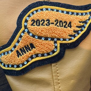 Op de afbeelding: Een aangepaste patch met een oranje vleugelontwerp, zwarte rand en sprankelende accenten. De patch heeft de naam "ANNA" en het jaar "2023-2024". De patch is genaaid op een bruin lederen oppervlak.