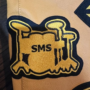 May include: A close-up of a tan leather jacket with embroidered patches. The main patch is a yellow drum set with the letters "SMS" in black. The patches are outlined in navy blue.