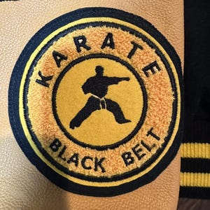 May include: Close-up of a tan and black jacket with a Karate Black Belt patch. The patch is circular with a yellow center, a black silhouette of a karate practitioner, and the words "KARATE BLACK BELT".