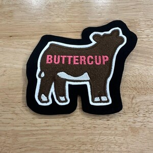 May include: An embroidered patch shaped like a cow with brown fur, white markings on the legs and belly, and a black border. The word BUTTERCUP is stitched in pink capital letters across the center of the cow's body against the brown background.