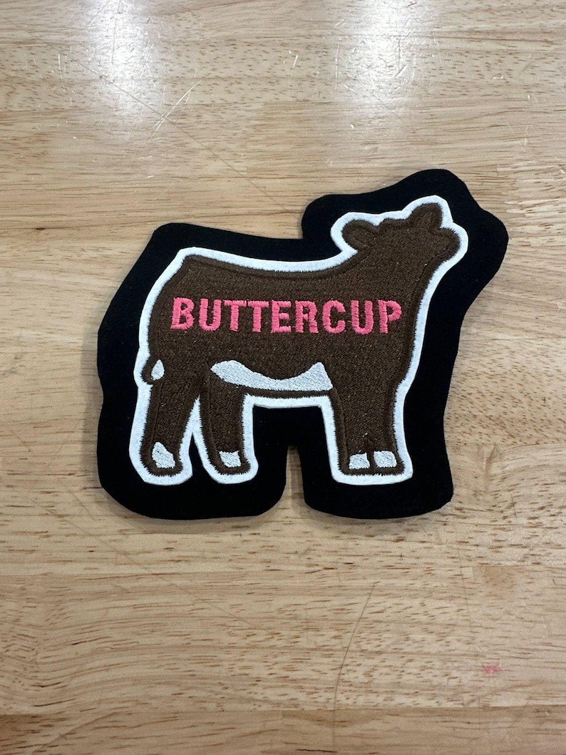 May include: An embroidered patch shaped like a cow with brown fur, white markings on the legs and belly, and a black border. The word BUTTERCUP is stitched in pink capital letters across the center of the cow's body against the brown background.