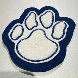 May include: A blue and white paw print patch. The patch features a white, textured paw print design with blue stitching and a blue border. This patch is likely for apparel or accessories.