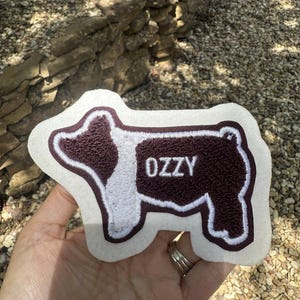 Puede incluir: Parche bordado con forma de oveja, con borde blanco y el nombre "OZZY" en letras blancas. El cuerpo de la oveja es de color granate oscuro.