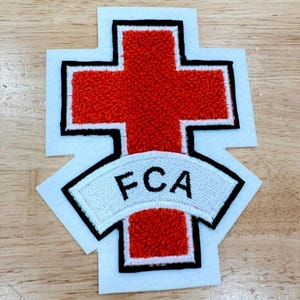May include: A patch featuring a red cross with a textured appearance, outlined in black. A white banner with the letters "FCA" is positioned below the cross. The patch has a white border and is likely for apparel or accessories.