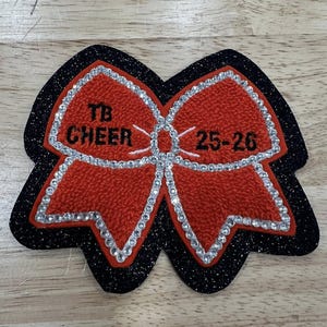 Może przedstawiać: Czerwono-czarna naszywka w kształcie kokardy do cheerleadingu. Kokarda ma czerwony, teksturowany środek z białym napisem "TB CHEER 25-26". Kokarda jest obrysowana srebrnymi dżetami i czarną, błyszczącą obwódką.