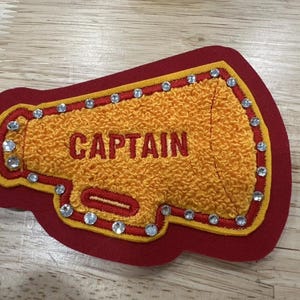 May include: A red, yellow, and orange megaphone-shaped patch with the word "CAPTAIN" embroidered in orange. The patch is outlined in yellow and red, with clear rhinestones around the edge.