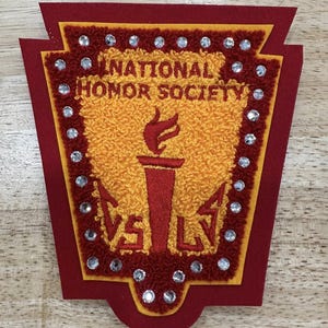 May include: A red, gold, and yellow felt patch with the text "NATIONAL HONOR SOCIETY" and a torch illustration. The patch is bordered with small, silver rhinestones and the letters "VS LV".