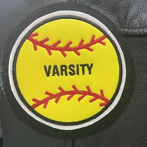 Peut inclure: Veste en cuir noire avec un écusson de softball jaune. L'écusson présente des coutures rouges et le mot "VARSITY" en lettres noires. L'écusson est entouré d'une bordure blanche.