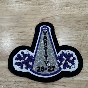 May include: A black, white, and purple Varsity cheerleading patch. The patch features a silver glitter megaphone with the word "VARSITY" and the year "26-27" in purple. Two purple and white pom-poms are on either side.