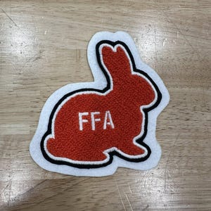 Puede incluir: Un parche de conejo FFA rojo y blanco. El conejo está delineado en blanco y negro, con las letras "FFA" bordadas en blanco sobre el cuerpo rojo. El parche es probablemente para un miembro de Future Farmers of America.