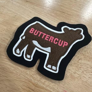 May include: A brown cow-shaped embroidered patch with a black border and white trim. The word BUTTERCUP is stitched in pink thread across the body of the cow. The patch features a white patch on the cow's belly and white markings on the legs.