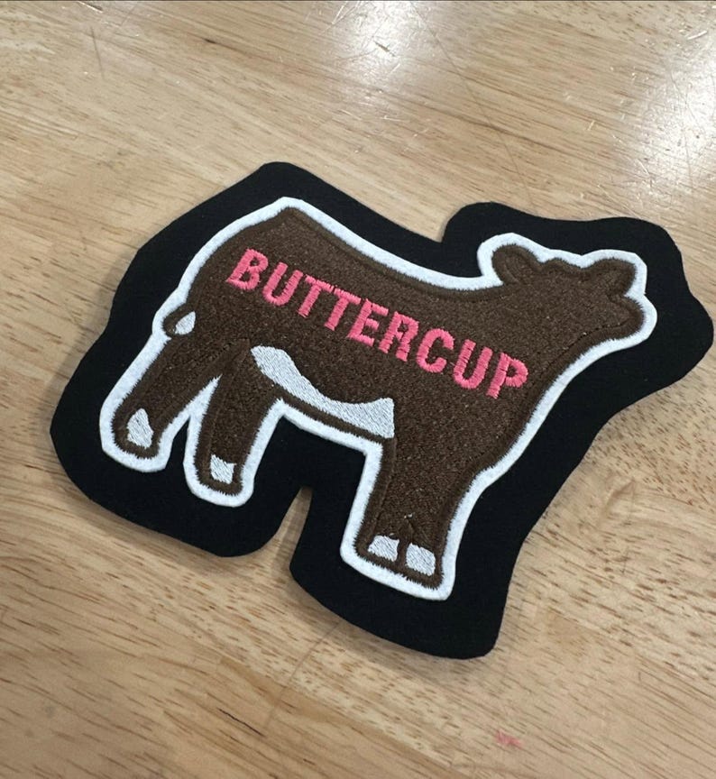 May include: A brown cow-shaped embroidered patch with a black border and white trim. The word BUTTERCUP is stitched in pink thread across the body of the cow. The patch features a white patch on the cow's belly and white markings on the legs.