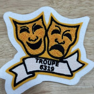 May include: Embroidered patch featuring two theater masks, one smiling and one frowning, in yellow and black. A white banner with the text "TROUPE 6319" is below the masks. The patch has a white border.