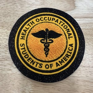 May include: A round patch with the text "HEALTH OCCUPATIONAL STUDENTS OF AMERICA" in yellow lettering on a black background. The center features a yellow textured circle with a black caduceus symbol.
