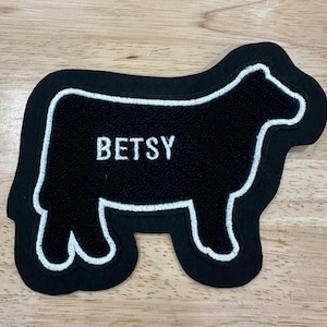 Puede incluir: Un parche negro con forma de oveja con ribete blanco y el nombre "BETSY" bordado en blanco. El parche tiene un borde negro y probablemente está hecho de una tela texturizada. El diseño es simple y limpio, adecuado para diversas aplicaciones.