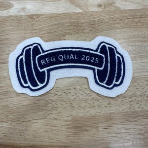 Puede incluir: Un parche de fieltro blanco con forma de mancuerna con detalles en azul marino. El diseño de la mancuerna tiene un contorno blanco. El texto "REG QUAL 2025" está en azul marino sobre una pancarta blanca.