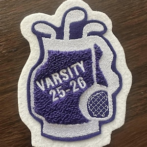 May include: A varsity patch in the shape of a golf bag, with purple embroidery and white trim. The text "VARSITY 25-26" is embroidered in white on the purple bag. The patch features golf clubs and a golf ball.