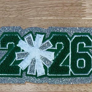 Könnte beinhalten: Ein Aufnäher mit den Ziffern 2 und 6 in Dunkelgrün mit strukturierter Oberfläche, getrennt durch ein weißes sternförmiges Symbol. Das gesamte Design ist umrandet und mit silbernem Glitzermaterial hinterlegt.