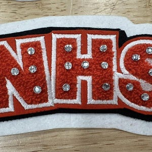 以下が含まれることがあります： 赤、白、黒の文字「NHS」が刺繍されたパッチ。文字はラインストーンで装飾され、質感のあるふわふわした外観です。パッチは白いフェルトの背景にあります。