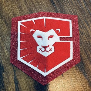 May include: A red and white embroidered patch with a lion's head design. The patch is shield-shaped with a glittery red border. The lion's head is white, and the background is red.