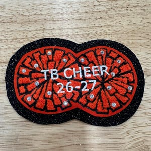 May include: A cheerleading patch in the shape of two pom-poms. The pom-poms are red with black outlines and a black glitter border. The text "TB CHEER 26-27" is in white. Rhinestones are scattered across the design.