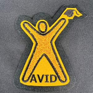 May include: A felt patch featuring a yellow figure with outstretched arms and a graduation cap. The figure is outlined in yellow with a black border. The word "AVID" is embroidered in yellow on the patch.