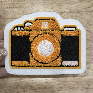 May include: Embroidered camera-shaped patch. The camera features a black body with orange and white detailing. The patch is edged with a white border.