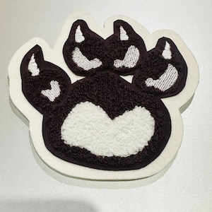 May include: An embroidered paw print patch in brown and white. The paw print has a textured, plush appearance with a white centre and white accents on the toes. The patch is outlined with a brown border and a white backing.