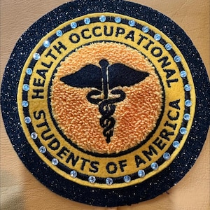 May include: A circular patch with the text "HEALTH OCCUPATIONAL STUDENTS OF AMERICA" in gold lettering. The patch has a black glitter border and a yellow center with a medical symbol. Rhinestones are placed around the border.