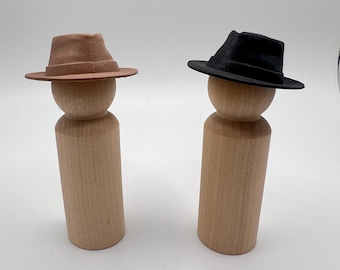 May include: Two light-coloured wooden peg dolls standing upright. The peg doll on the left wears a tan fedora-style hat, while the peg doll on the right wears a black fedora-style hat. Both figures have a smooth, cylindrical body and a rounded head.