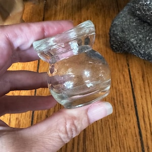 Puede incluir: Un pequeño tarro de vidrio transparente con un cuerpo redondeado y una abertura acampanada e inclinada. El tarro se sostiene en una mano, con una escultura de mano de madera y un mortero y maja de piedra gris oscuro en el fondo. El tarro parece ser vintage.