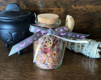 Puede incluir: Un frasco de vidrio lleno de sales de baño rosas y naranjas, atado con una cinta morada con un amuleto de pentagrama. El frasco está sentado sobre una superficie de madera con una cuchara de madera y un manojo de salvia.