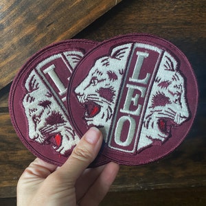 May include: Two maroon felt patches with white embroidered lion heads and the word "LEO" in white. The patches are sewn together and held in a hand.