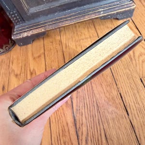 May include: A hardcover book with a dark cover and light-colored pages. The book's spine is visible, showing a textured gray binding. The pages are a light tan color. The book is held in a hand.