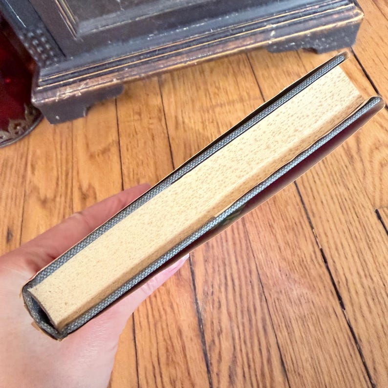 May include: A hardcover book with a dark cover and light-colored pages. The book's spine is visible, showing a textured gray binding. The pages are a light tan color. The book is held in a hand.