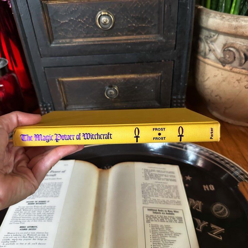 K&ouml;nnte beinhalten: Ein gelbes Buch mit dem Titel "The Magic Power of Witchcraft" von Frost. Das Buch hat ein violettes Ankh-Symbol auf dem Cover.