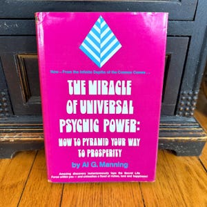 May include: A vintage book titled "The Miracle of Universal Psychic Power" by Al G. Manning. The book has a bright pink cover with white text and a blue and white geometric design. The book's cover also includes additional text.